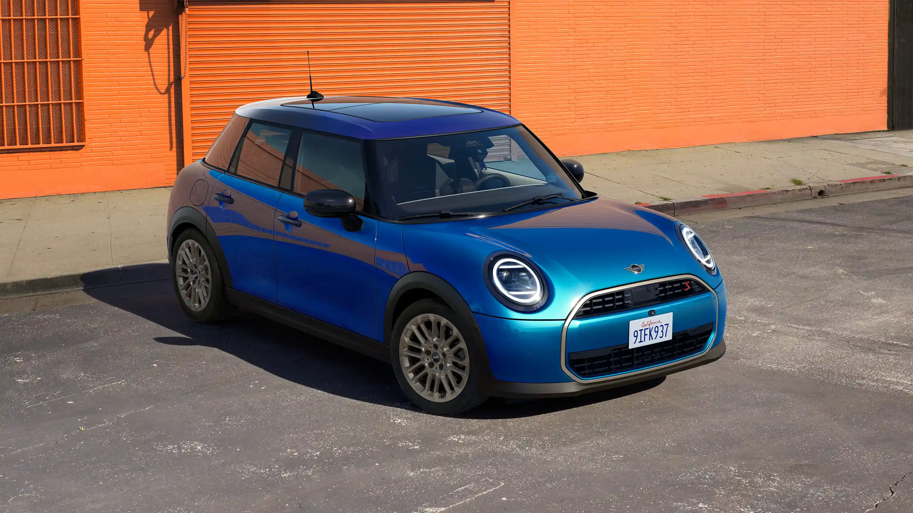 Frontal view of a parking MINI Cooper 5-Door in Icy Sunshine Blue and Multitone Blue Roof on street with orange wall in the background